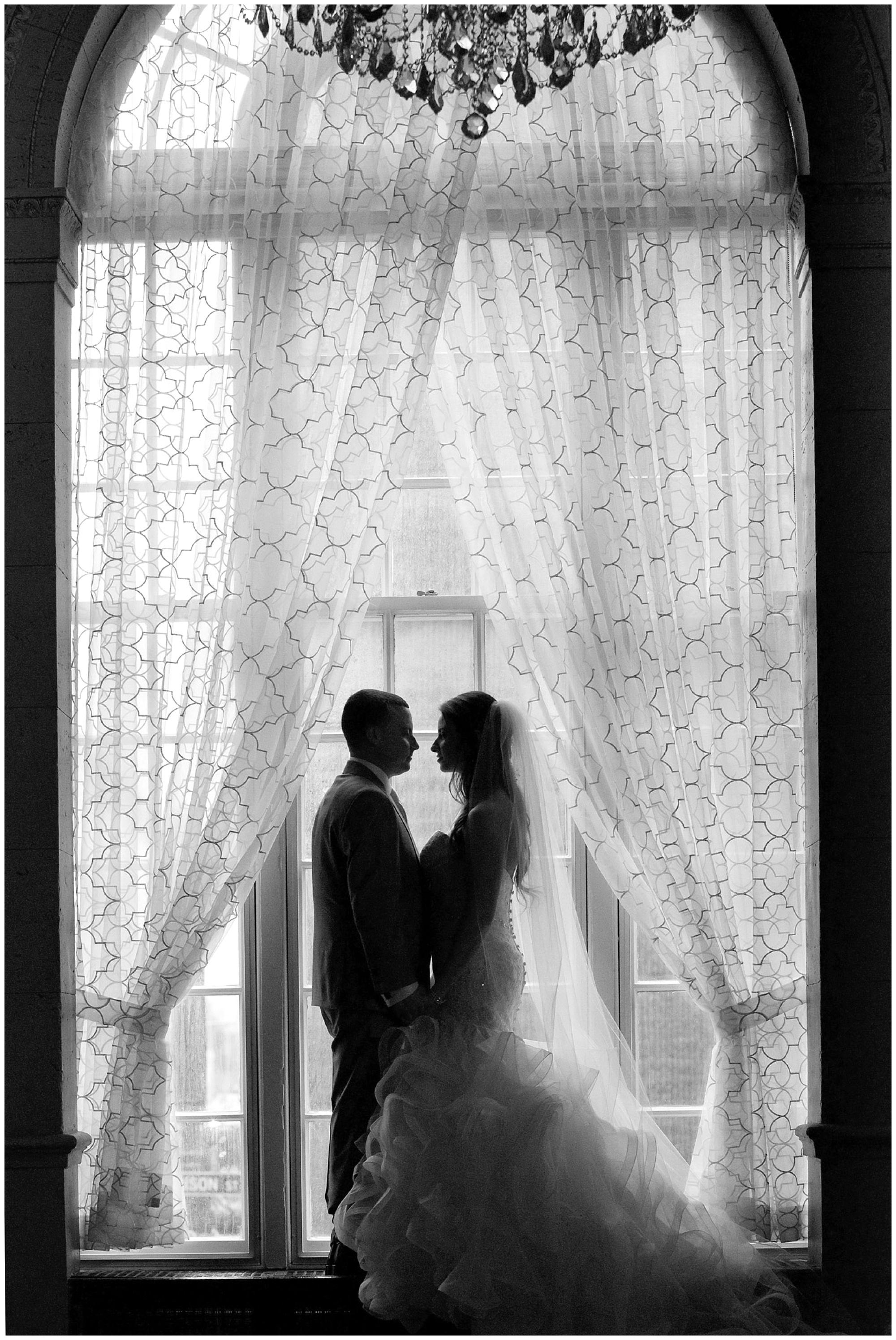 A portrait of a bride and groom semi silhouetted and framed by a large arched window.