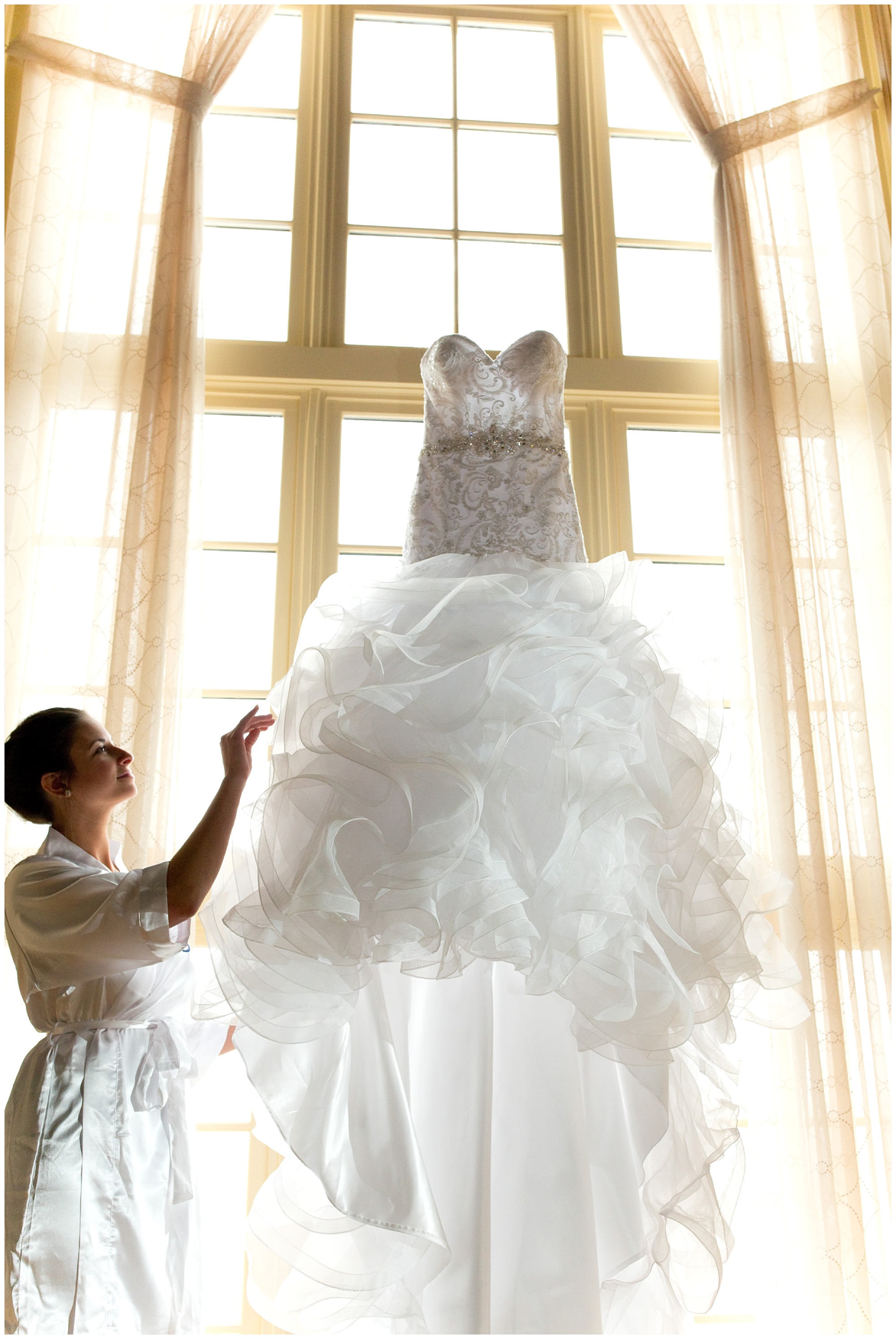 Photo of a bride admiring her dress which is suspended and backlited by a large window.