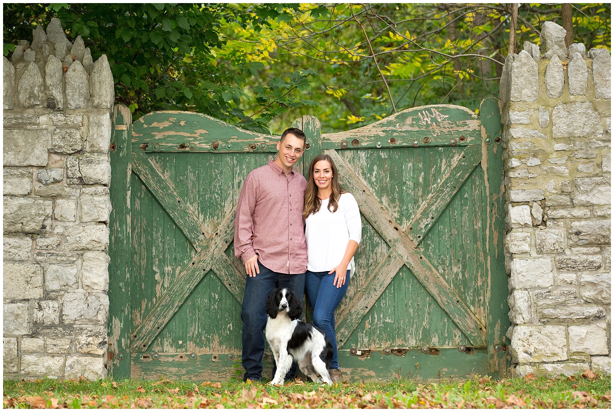 Photo of an engaged couple standing in front of a distressed green wooden gateway with their dog.