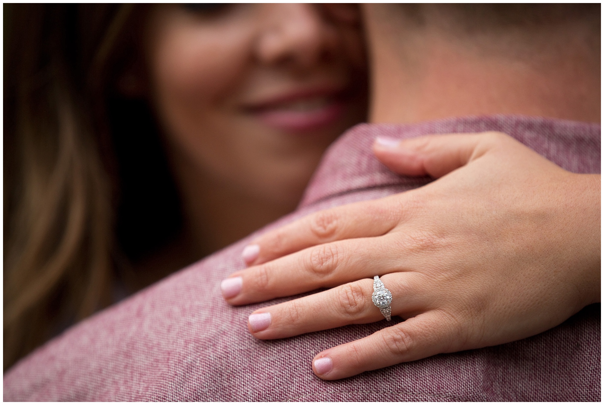 Photo of an engaged couple embracing with focus placed upon her engagment ring which is on his back..