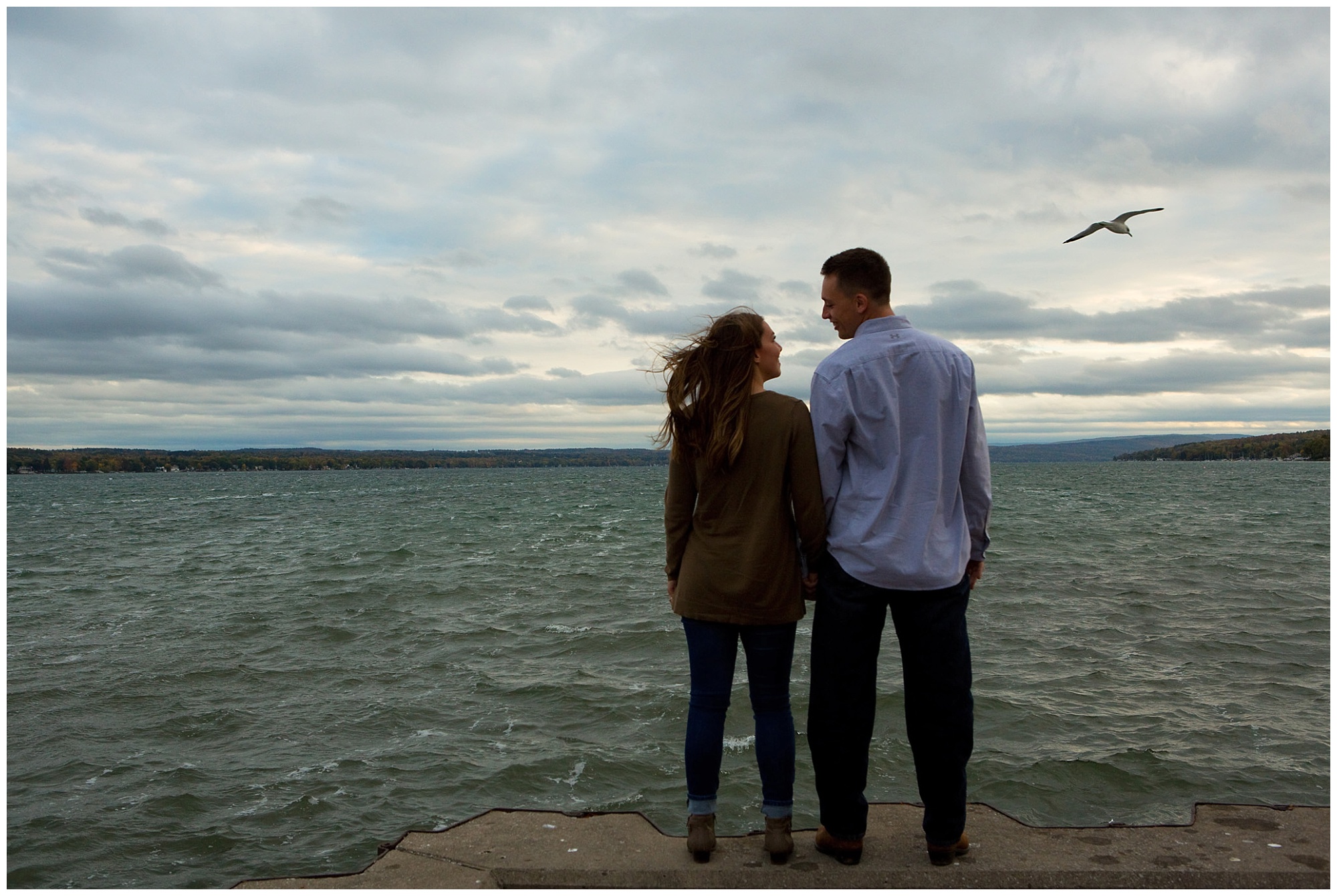 Photo of an engaged couple with thier backs to the camera looking at eachother with Canandaigua lake in the background
