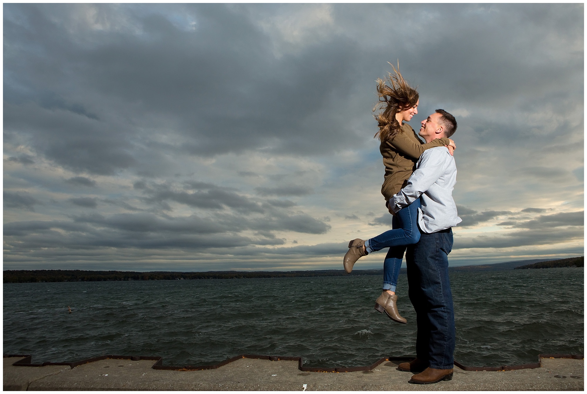 Photo of an engaged couple with her jumping into his arms with Canandaigua lake in the background.