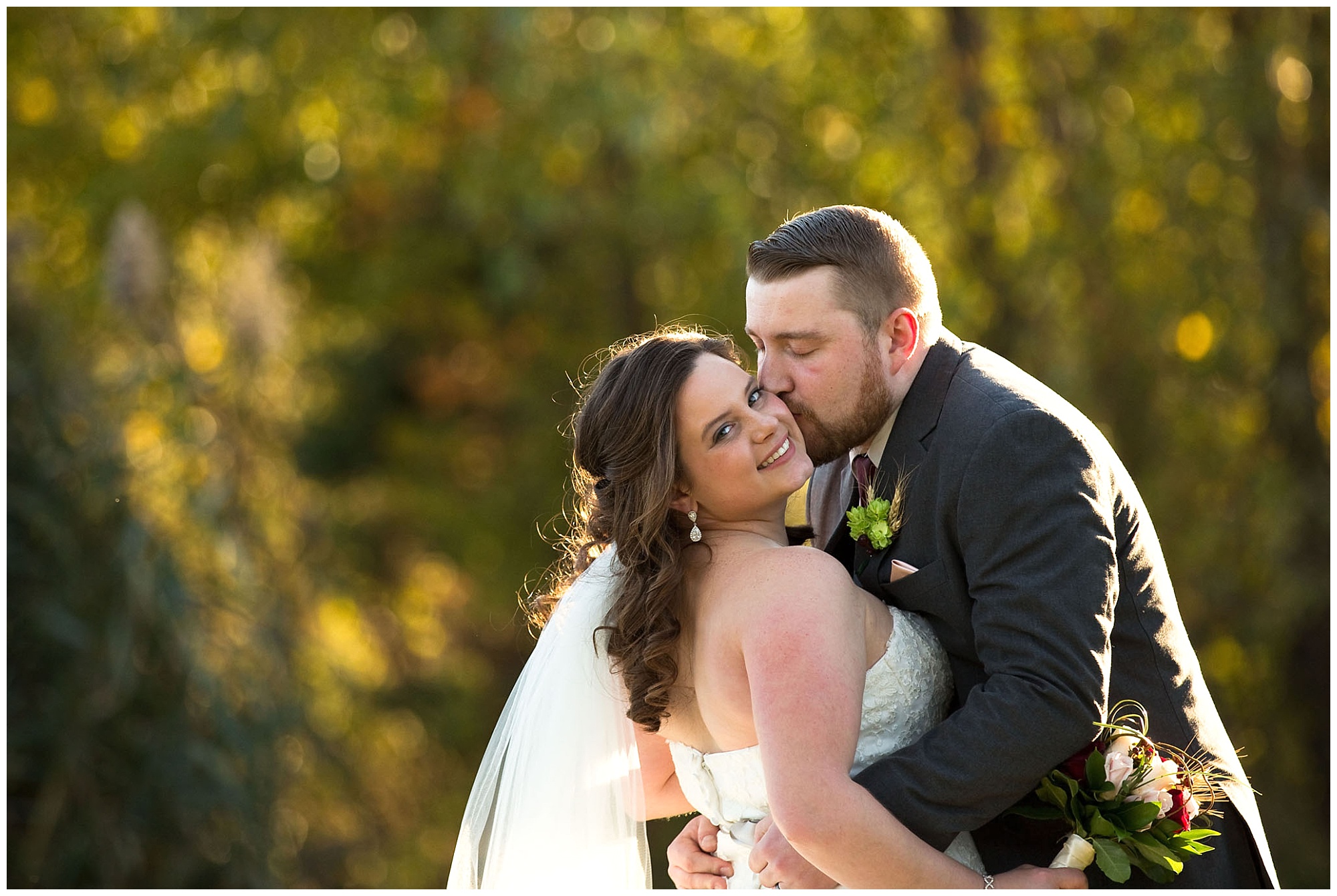 A photo of the groom kissing his bride while she leans back slightly looking back at the camera lens while backlighted by the late autumn afternoon sun.
