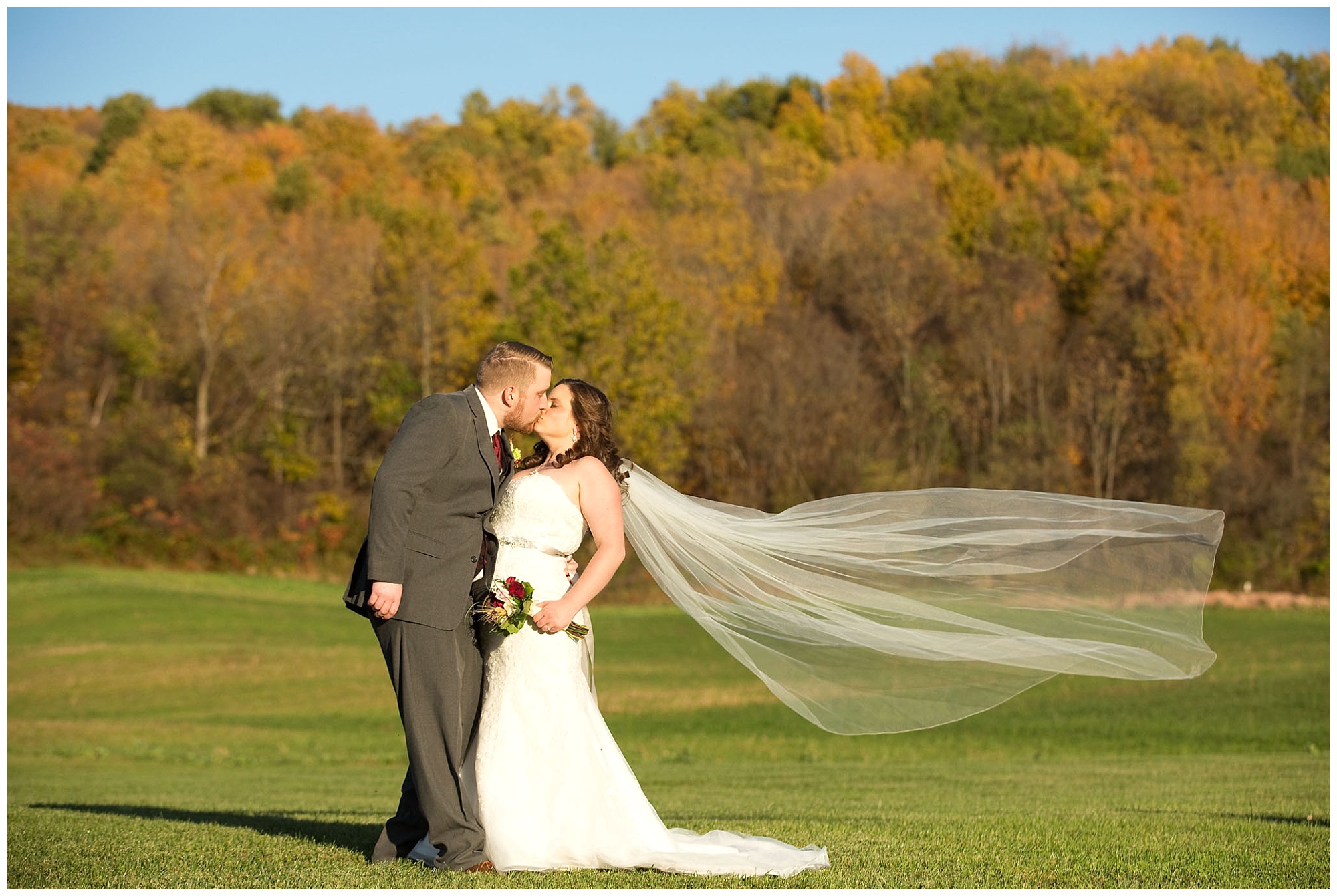 A bride and groom portrait in the late afternon sunlight with him kissinig her while her vails blows in the late autumn breeze.