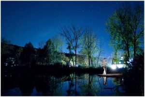 Wide photo of the bride and groom kissing under the stars by the pond.
