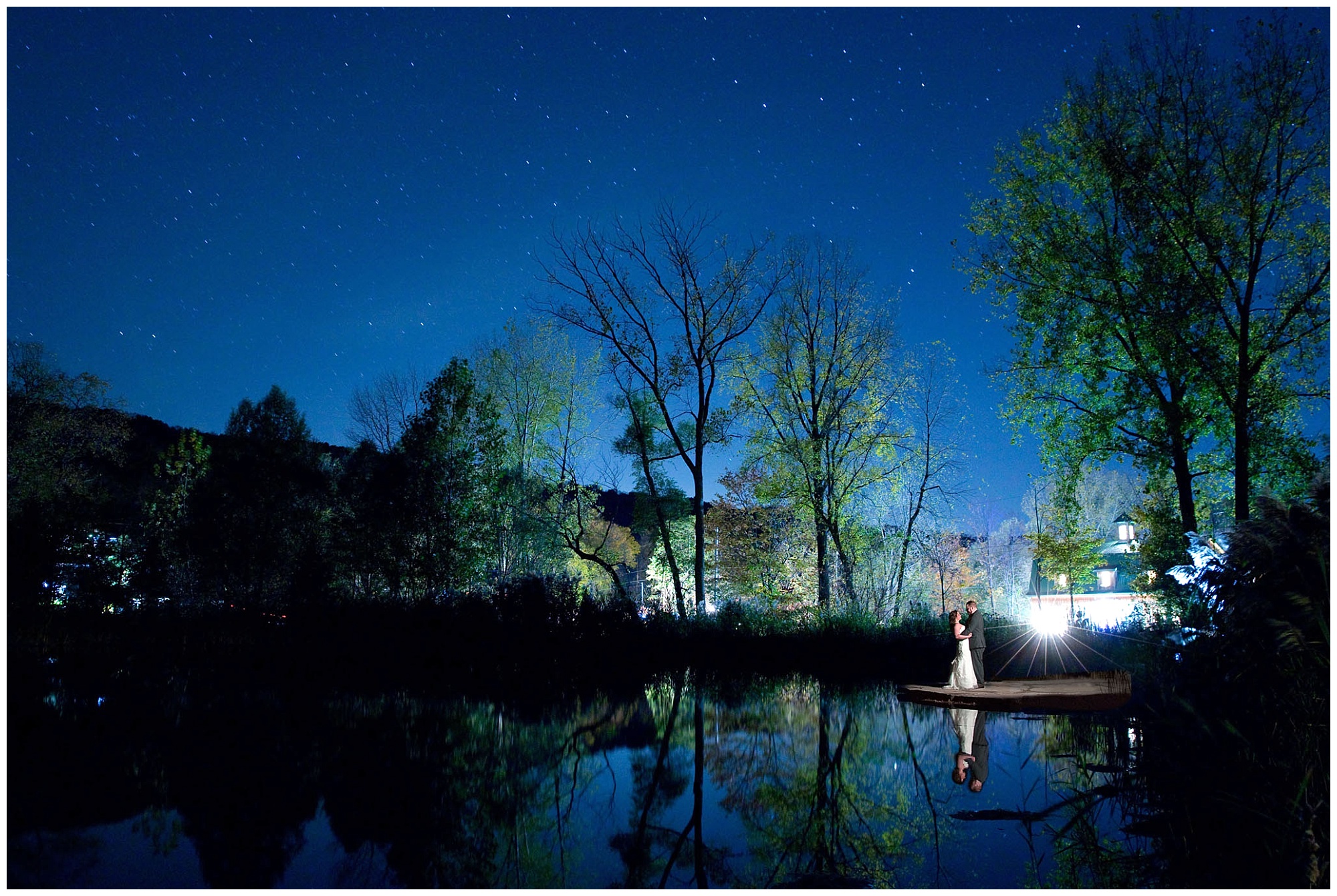 Wide photo of the bride and groom kissing under the stars by the pond.