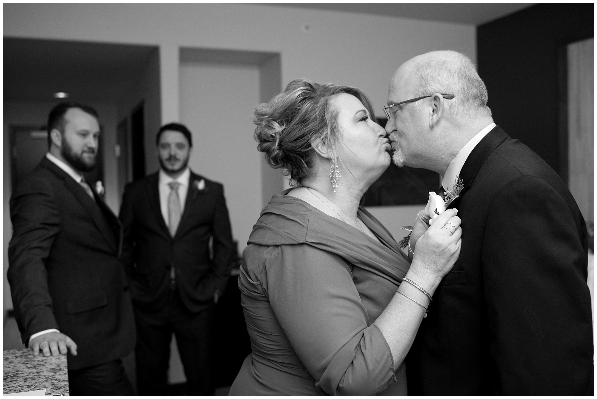 A photo of a groom's parents kissing with he and his best man in the background.