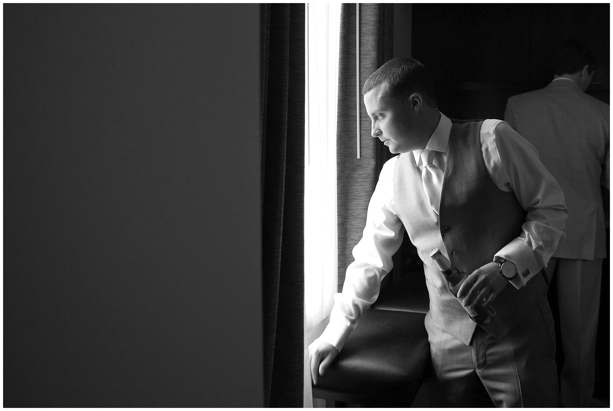 Photo of a groom peering out a window with a beer in his hand.